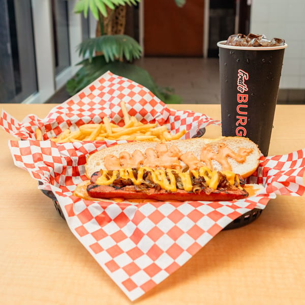 A Fred's Burger hot dog with cheese and onions, fries, and a soda on a wooden table.