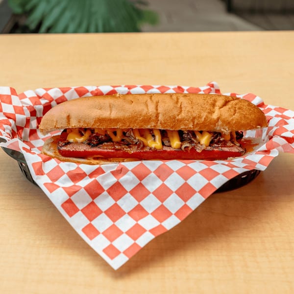A Fred's Burger hot dog with cheese and caramelized onions in a basket with red and white checkered paper.